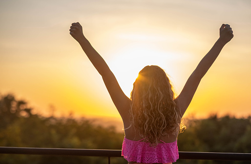 girl outside with raised arms looking at the sunset on the terrace with curly hair and in a bathing suit