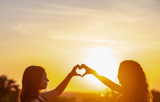 two happy girls outdoors at sunset making a heart shape together smiling