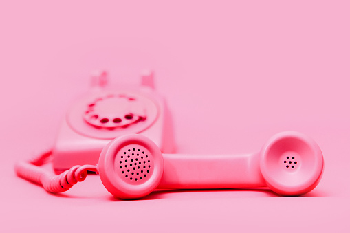 A pink old fashioned rotary style phone, photographed on a matching pink colored backdrop for a monochromatic style.  The receiver is off the hook.