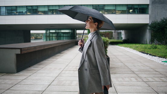 Elegant woman walking rainy day holding umbrella. Confident businesswoman stroll financial district checking smartphone. Relaxed office worker rest taking break outdoors. Corporate manager go meeting