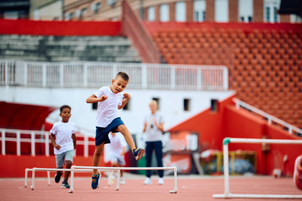 istock Happy kid jumping over obstacles while running on a track at the stadium. 1908079359