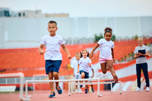 Group of kids jumping over obstacles while running during exercise class at the stadium. Focus is on African American girl.