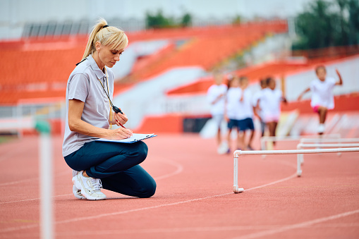 Athletics coach writing on clipboard during sports training at the stadium. The kids are in the background. Copy space.