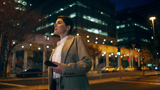 Woman waiting night taxi in downtown. Successful businesswoman hold mobile phone looking city road. Corporate manager commute checking cab service application online in evening. City lifestyle concept