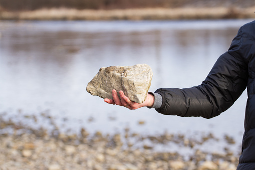 Мужская рука держит большой белый камень на фоне горной реки зимой.