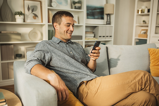 Shot of a happy young man using a mobile phone on the sofa at home