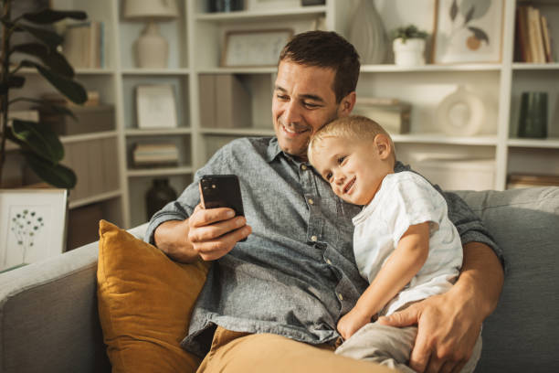 Modern fatherhood Father and son sitting on sofa and watching favorite cartoon on mobile phone. father holding son stock pictures, royalty-free photos & images