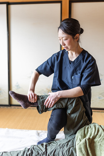 A Japanese female massage therapist stretches and massages a womans leg on a tatami floor of a traditional Japanese room.