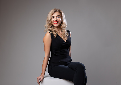 A portrait of a confident woman with blonde hair, wearing a black turtleneck and a pearl necklace. She has a bright smile and is posing against a grey background. The lighting accentuates her cheerful expression and elegant attire.