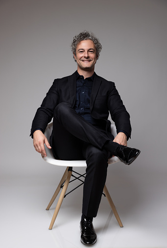 This image captures a portrait of a mature man with a confident and approachable smile. His curly gray hair adds a distinguished touch to his appearance. He is dressed in business casual attire, featuring a well-fitted black blazer and a dark shirt, conveying a sense of casual elegance. The subject is seated with his hands clasped, exuding an aura of positivity and professionalism. The neutral studio background ensures that the focus remains on him. This image would be well-suited for content related to business, leadership, lifestyle.