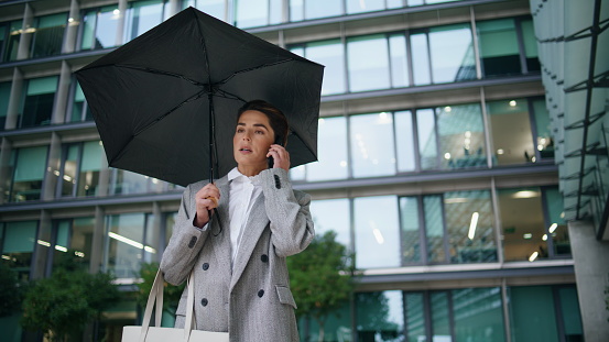 Corporate woman talk mobile phone at contemporary office building on rainy day. Stylish middle aged businesswoman hold umbrella speaking partner on street. Elegant manager making arrangements on call