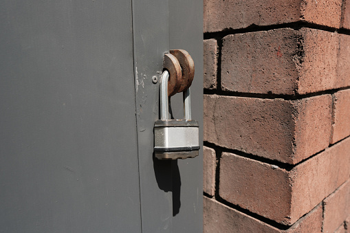 Close-up of a silver colored padlock securing a gray door next to a brick wall.