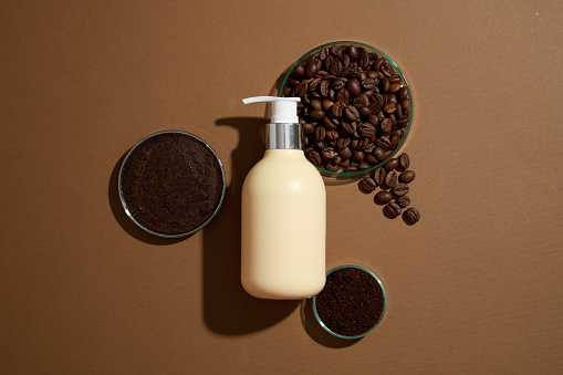 On a brown background, a pump cosmetic bottle displayed with petri dish filled with coffee beans and coffee grounds. Top view, mockup scene for advertising product for body care