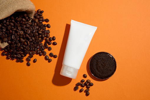 A white plastic tube displayed on orange background with coffee beans and coffee grounds. Mockup scene for advertising. Coffee grounds with countless tiny particles help gently remove dead skin cells