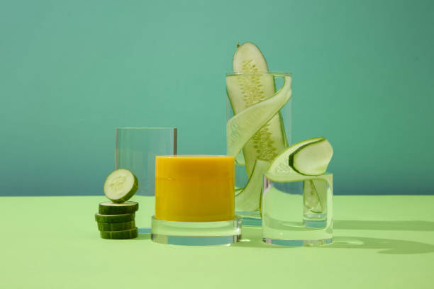 istock Front view of an unlabeled yellow cosmetic jar placed on a glass platform, surrounded by glasses with thin slices of cucumber on a blue background. 1908573070