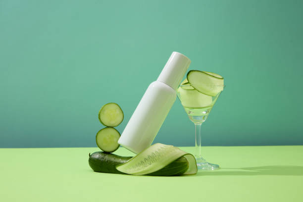 istock Thin slices of cucumber inside a cocktail glass, a white cosmetics bottle and fresh cucumbers displayed against a blue background. Blank label for letter design for branding. 1908573992
