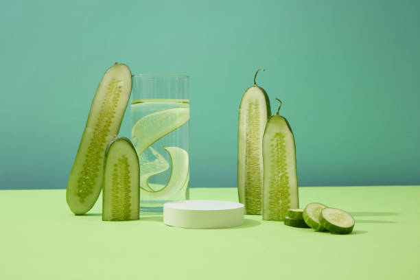 istock Cucumber slices inside a clear glass of water, surrounded by fresh cucumbers and an empty platform. Minimalist space for product display with blue background. 1908575138