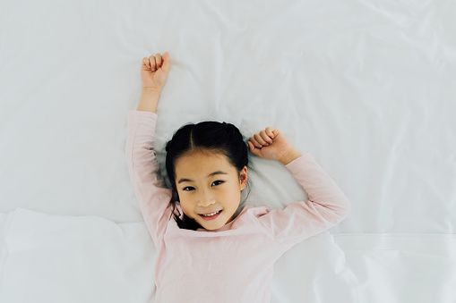 Portrait of young happy and cheerful Asian girl lying on back smiling on bed. She's looking at camera in casual clothing.