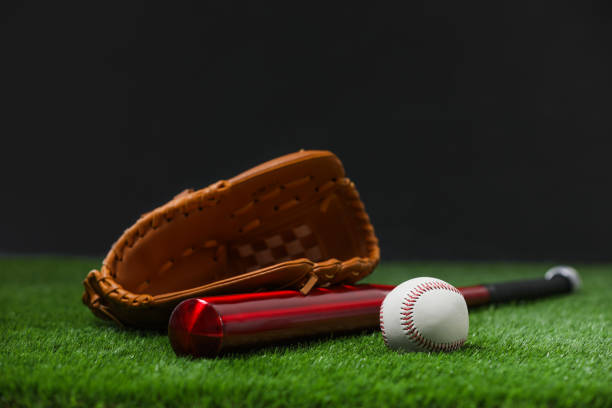 istock Baseball bat, leather glove and ball on green grass against dark background. Space for text 1908631368