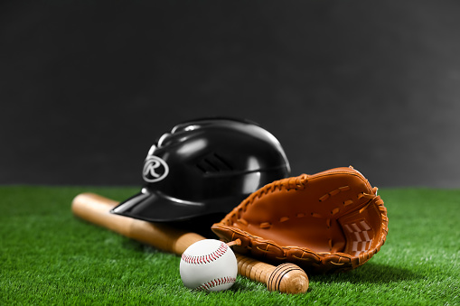Baseball bat, batting helmet, leather glove and ball on green grass against dark background