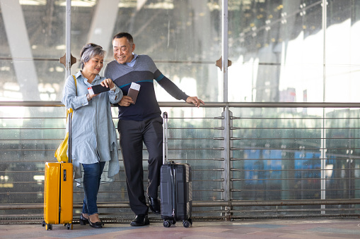 Portrait of Happy Asian senior couple holding passport and boarding pass walking together to airline check in counter in airport terminal. Elderly people travel by air transportation.