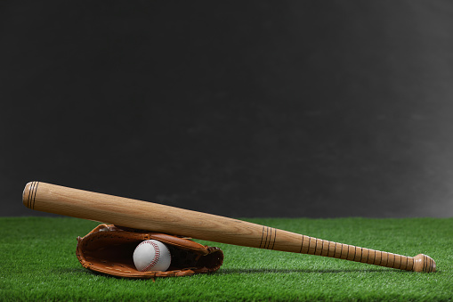 Baseball bat, leather glove and ball on green grass against dark background. Space for text