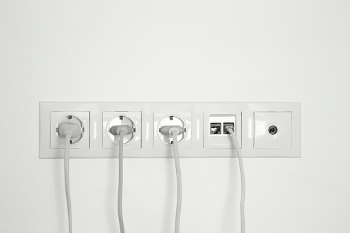 Many power sockets with plugs, ethernet and TV coax plates on white wall indoors