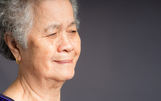Portrait of an elderly Asian woman with short gray hair looking away with a smile while standing on gray background in the studio. Space for text. Aged people and relaxation concept