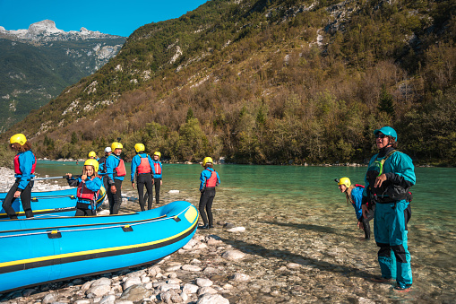 Ready for the thrill, a diverse group clad in safety gear stands by their raft at the edge of a clear river, mountains rising in the distance.