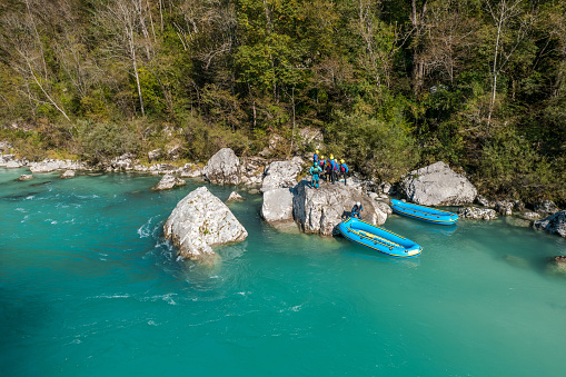 A group of rafters in blue and red life jackets takes a break on the rocky banks of a clear, flowing river surrounded by lush greenery.