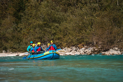 A Caucasian mother and her mixed-race children, one boy and two girls, navigate turquoise river waters with a professional guide, all clad in safety gear and vibrant life jackets.