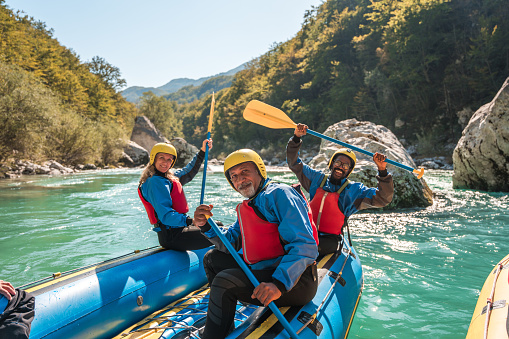 Mature men and women in protective rafting attire, with life vests and helmets, enjoying a group river rafting adventure on a scenic river.