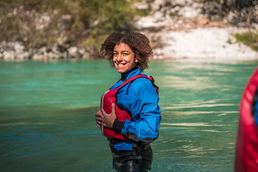 Mixed race young female in blue and red rafting gear with a yellow helmet holds an oar, displaying readiness for water sports in a natural setting.