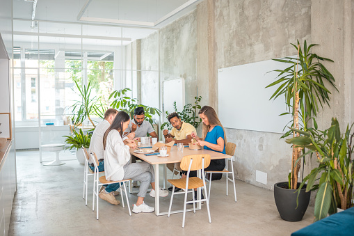 Colleagues with diverse looks enjoying lunch together in a stylish office setting. Seated and bonding over takeout, they establish a laid-back atmosphere.