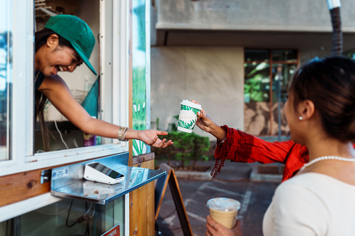 A smiling multiracial barista stands inside a food truck, leaning out the window and handing a to-go coffee to a group of customers.