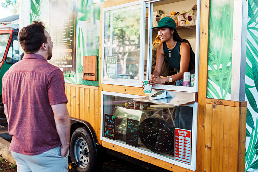 A male customer of Caucasian ethnicity stands outside smiling as he receives a drink from an unrecognizable female barista standing in a food truck.