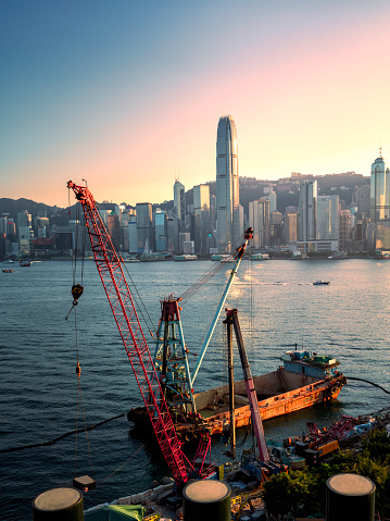 A pile driver, crane, and ship crane are operating on the waterfront in the West Kowloon Cultural District with the background of center of Hong Kong Island.