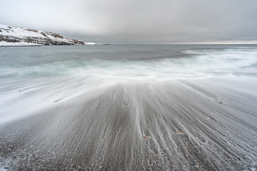 Tranquil ocean waves crash against sandy shore under cloudy sky. Ekkerøya, Northern Norway
