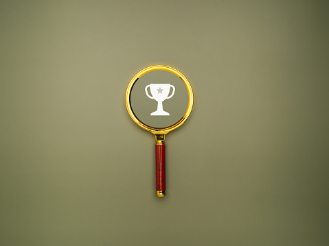 A magnifying glass with a white trophy on a green background. Top view. Flat lay. Ranking for winner business or sports competition. Concept of success, winner, victory, or top ranking