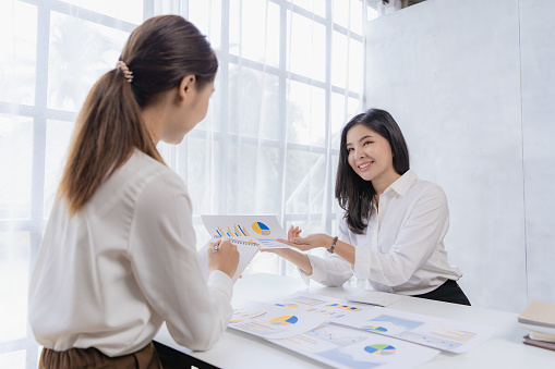 Two Asian female accountants have team meeting to summarize financial information in business office, creative female executive using tablet PC and smiling