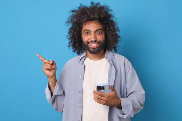 istock Young happy Arabian man with mobile phone points finger to side stands in studio 1908874814
