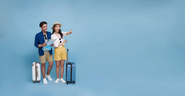 istock Happy Asian couple tourist looking map and hand pointing to copy space with baggage going to travel on holidays isolated on blue panorama background. 1908882433