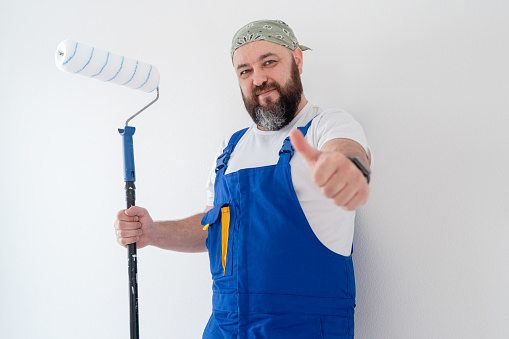 A positive man with a paint roller at the white wall, looking at camera and thumbs up gesturing