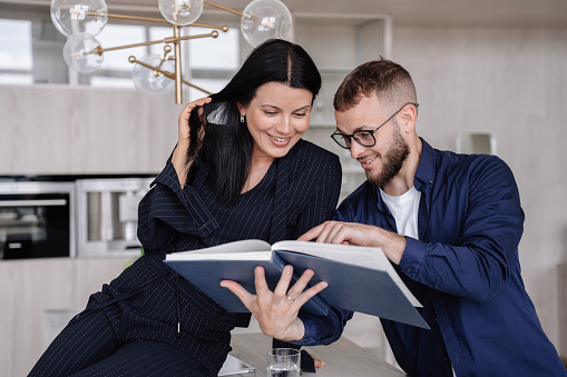 Young caucasian couple at home watching furniture catalog  choosing bed for new apartment, Newlyweds holding together wedding book, anniversary. Interior designer offering solutions for living room.