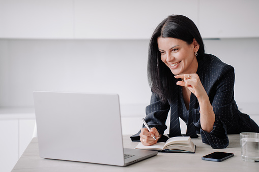 Successful brunette businesswoman in dark suit sitting at desk using laptop makes video call shows small size gesture by fingers. Cheerful European female lawyer remote works home. Career, finance.
