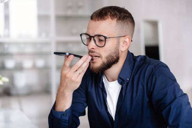 il giovane barbuto serio in occhiali registra il messaggio audio si siede a casa. l'uomo d'affari italiano in camicia blu parla al telefono usando il microfono. persone finanziarie, impiegato, manager. agente immobiliare professionista. - domestic staff audio foto e immagini stock