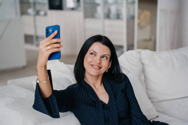 istock Smiling woman in a chic pinstripe suit taking a selfie on a comfortable sofa at home 1908952151