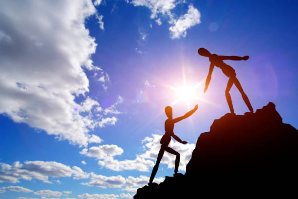 istock Two silhouettes mannequin climb mountain, one helping other, extending helping hand. Against background sky, sun and clouds. Concept of help and success in teamwork. 1908960562