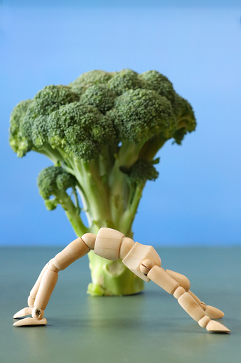 Stock photo showing close-up view of a wooden lay figure, jointed artist's model doing backbend yoga wheel pose in front of tree-like stem of broccoli pictured against a blue background bearing a resemblance to the sky.
