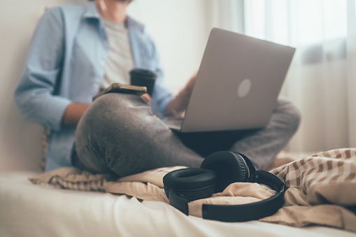 Focus on headphone. People sitting on bed holding coffee glass and using computer laptop. Freelancer working online at home. Meeting video conference with customer or connecting with social network